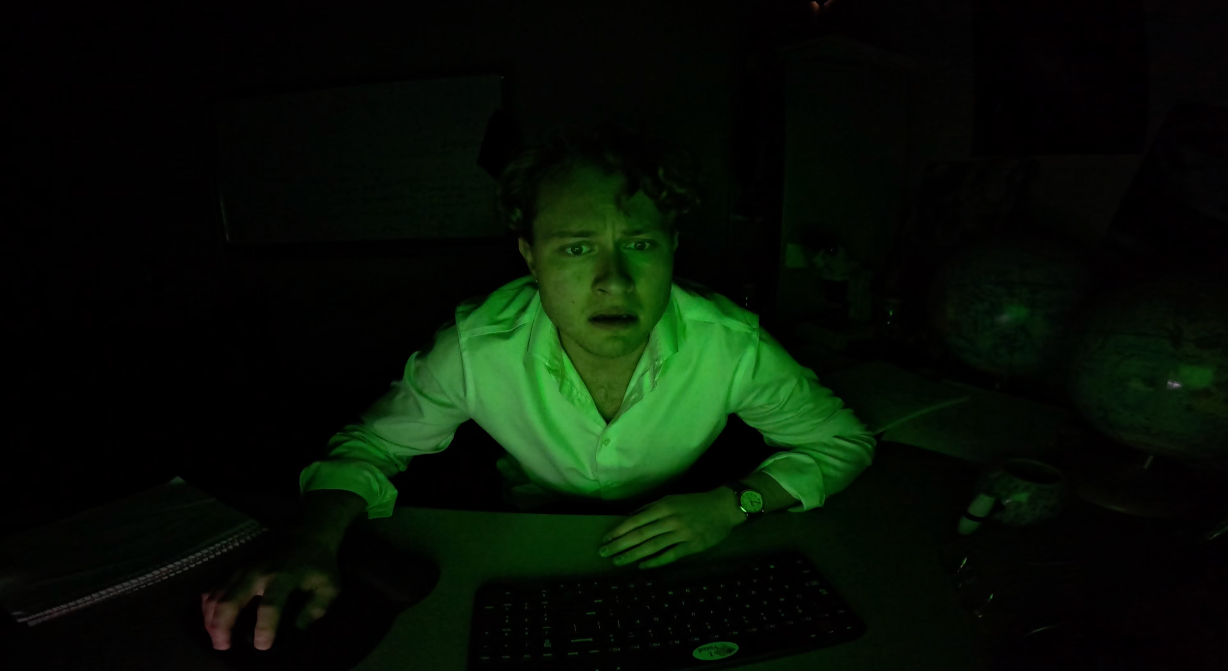 Jacob Leo Marino in NAME (2026), wearing a white shirt appears surprised, sitting at a desk with a computer keyboard, mouse, and notebook. The room is dark, illuminated by green light.