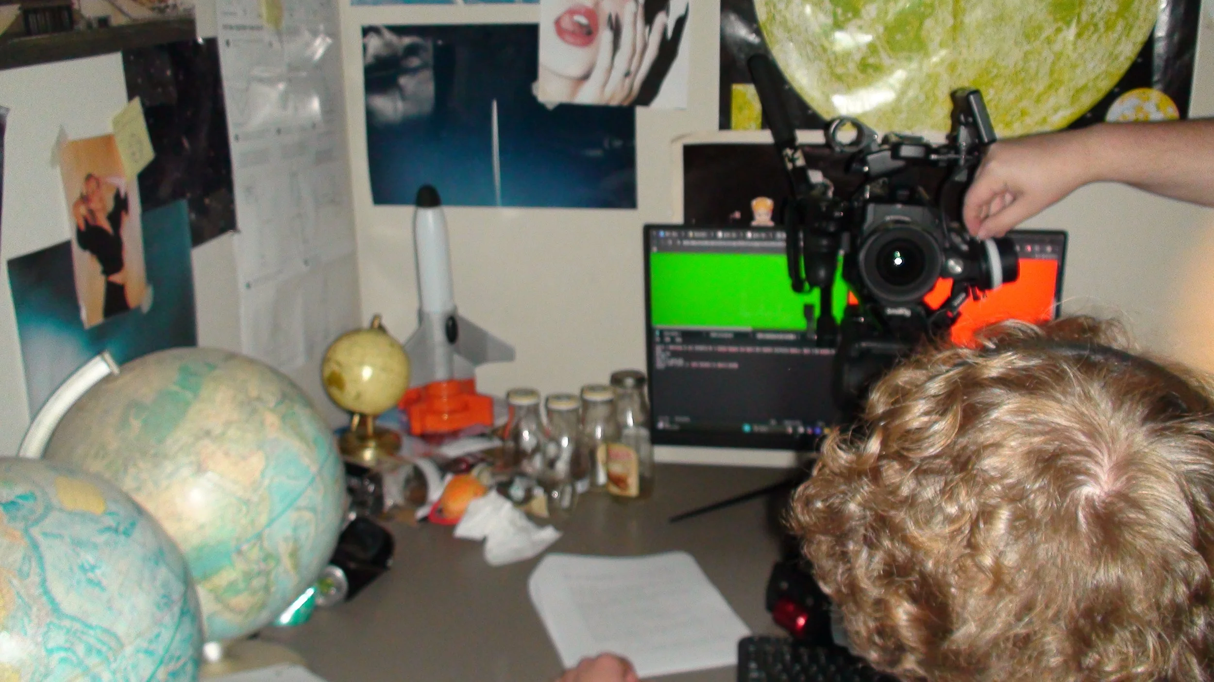 Person operating a camera on a desk with globes, bottles, a rocket model, and a monitor displaying green and red screens. BTS from the short film NAME (2026).