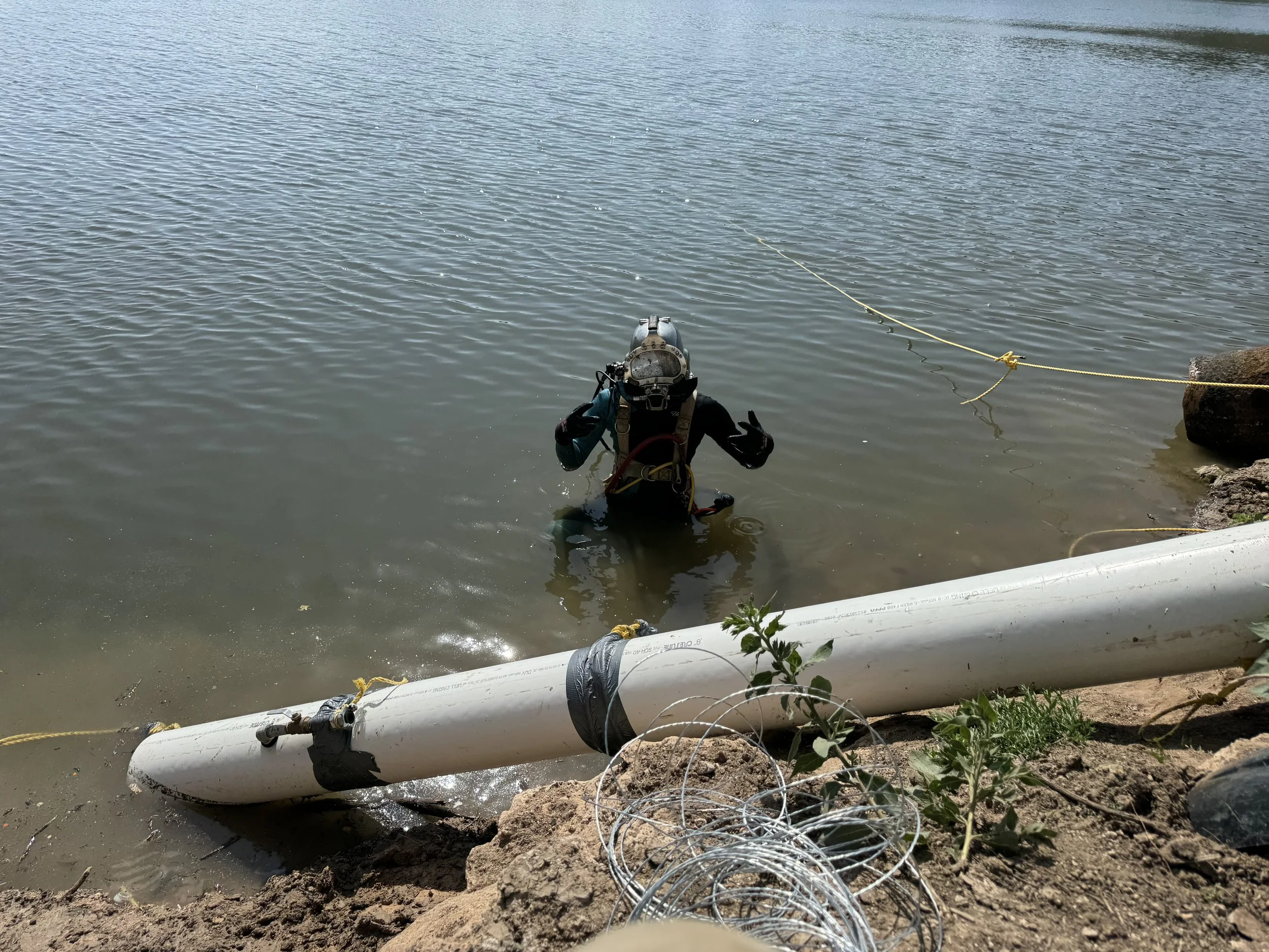 A diver in a wetsuit and helmet wades into a body of water, giving a peace sign with their hand. A large white pipe is partially submerged on the shore, with a coil of wire nearby and a yellow rope securing the pipe to a rock.