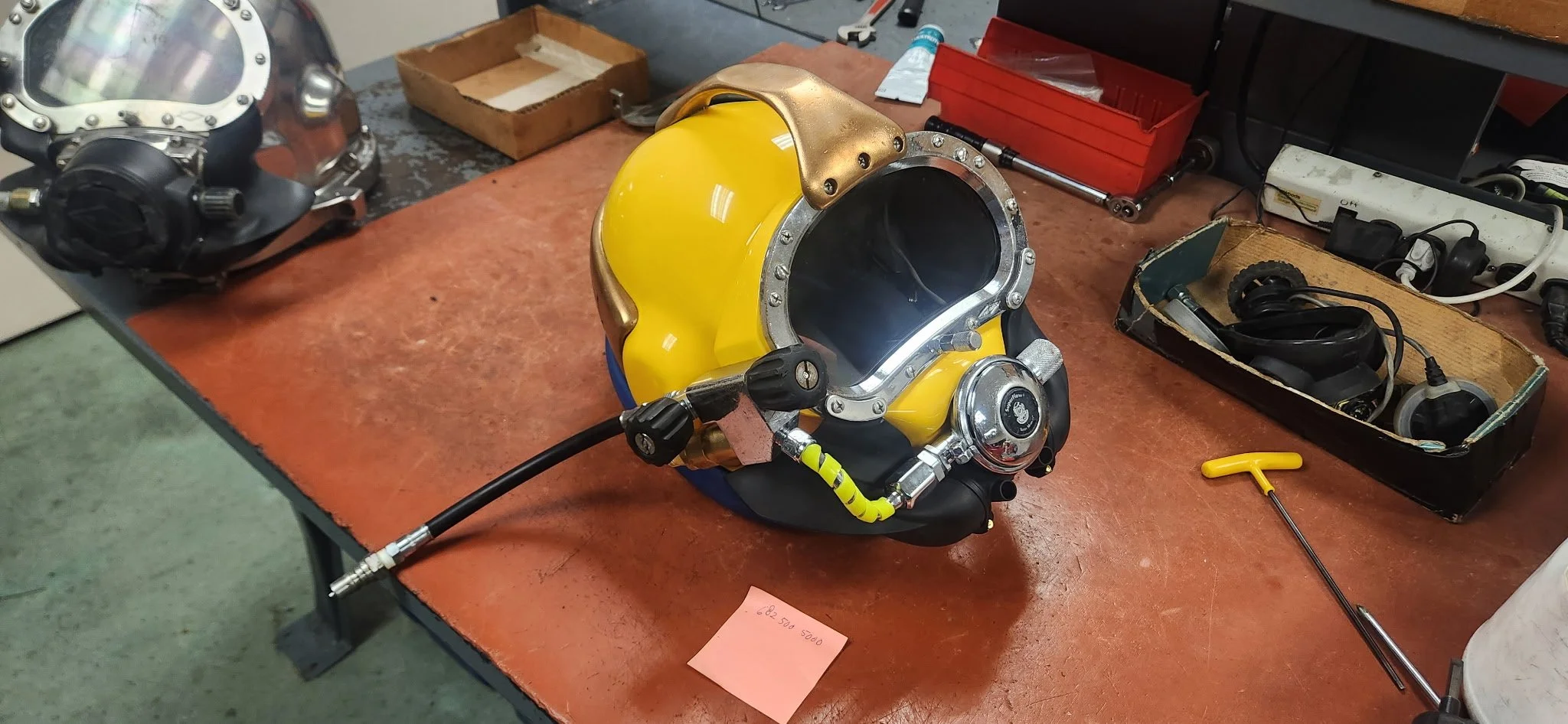 A yellow diving helmet on a workbench surrounded by tools and equipment, with a black mask to the left and organized boxes and cords in the background.