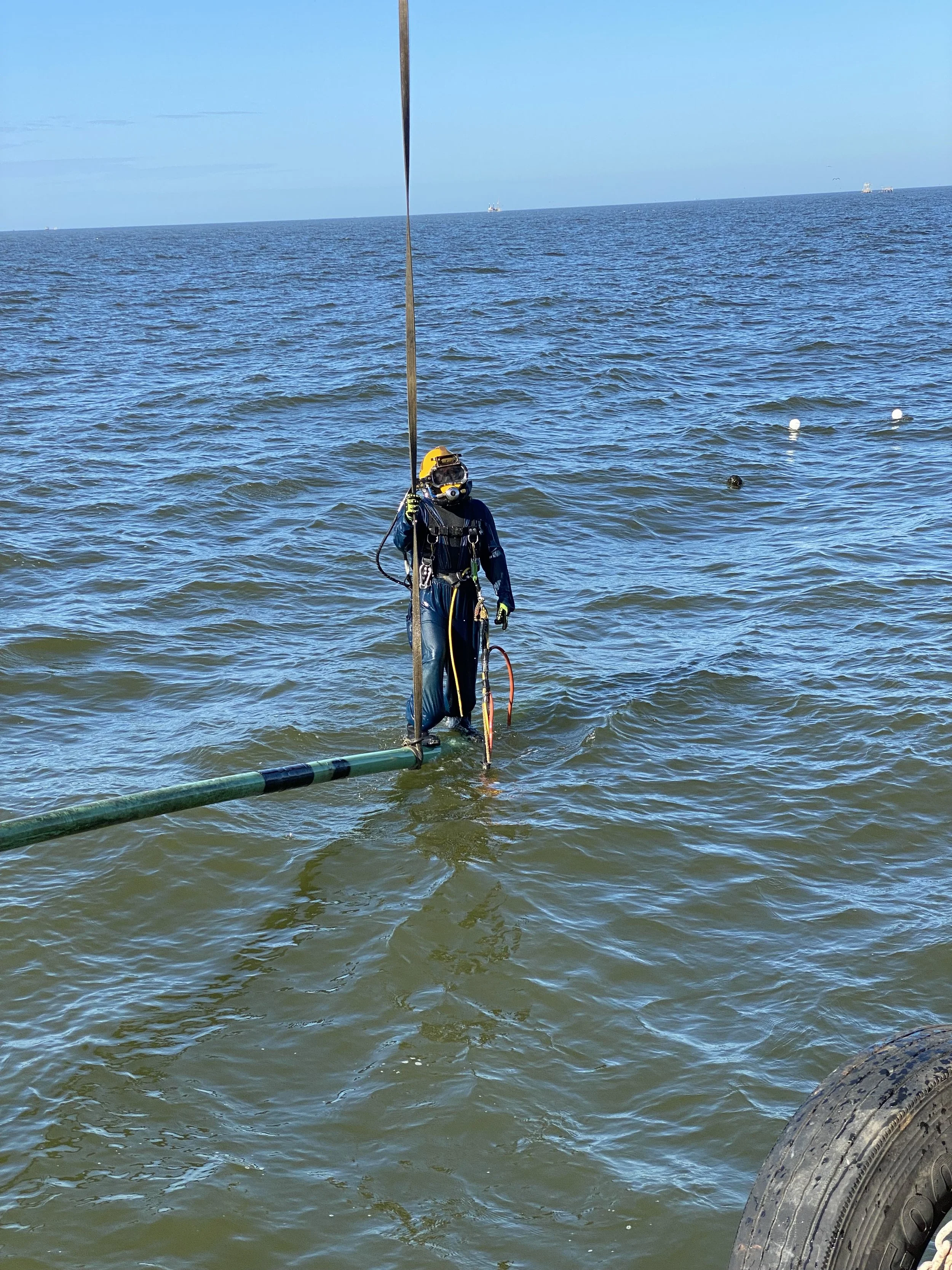 A scuba diver in a wetsuit stands in shallow water holding a pole with an underwater pipe or cable, with the ocean and a clear blue sky in the background.