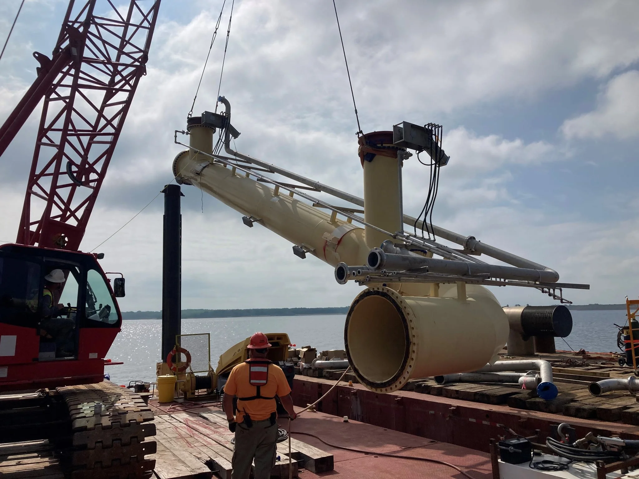 Construction workers operate a crane on a barge, installing large industrial pipes with a body of water in the background.
