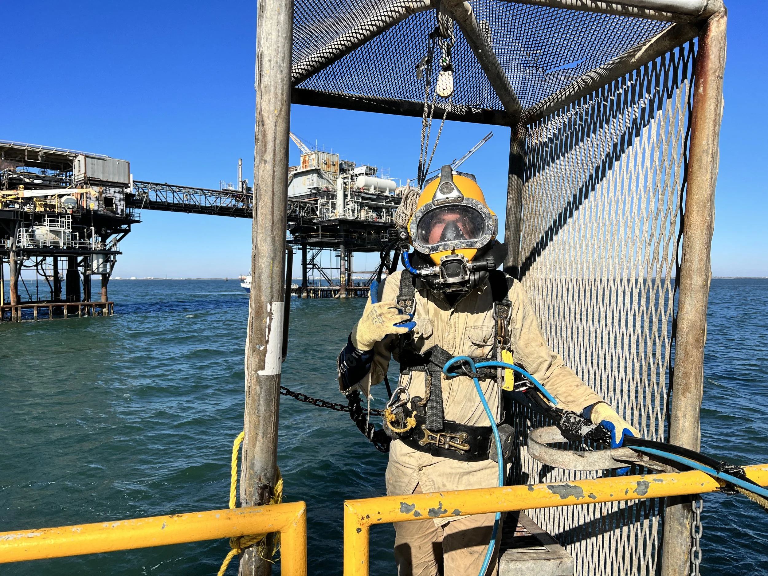 A diver wearing a yellow helmet and wetsuit standing on a metal platform with a safety railing, near an offshore oil rig, making a hand gesture with one hand, holding equipment with the other, with blue water and a clear sky in the background.