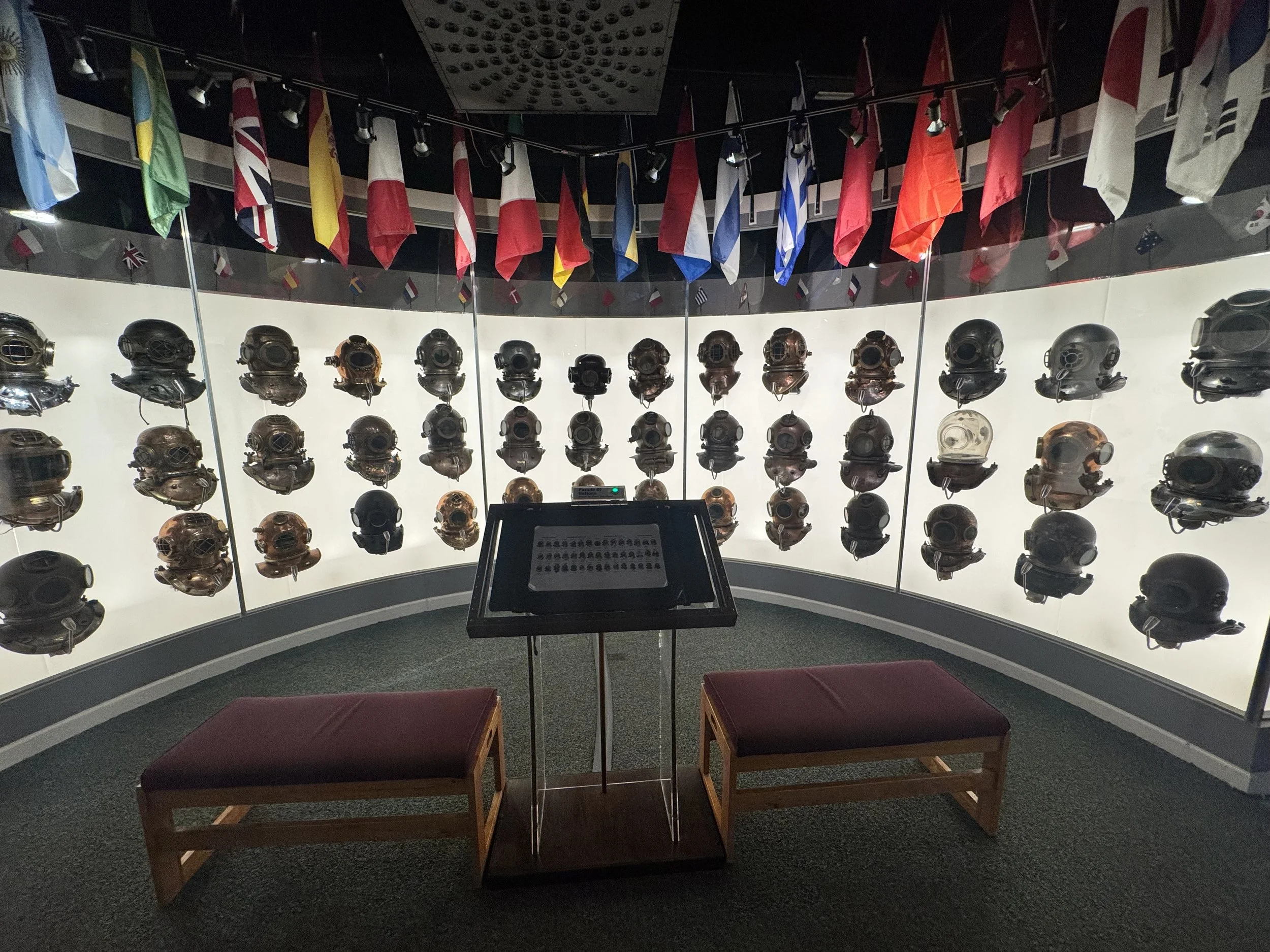 A display of vintage diving helmets on a curved backlit wall at a museum, with international flags hanging above.