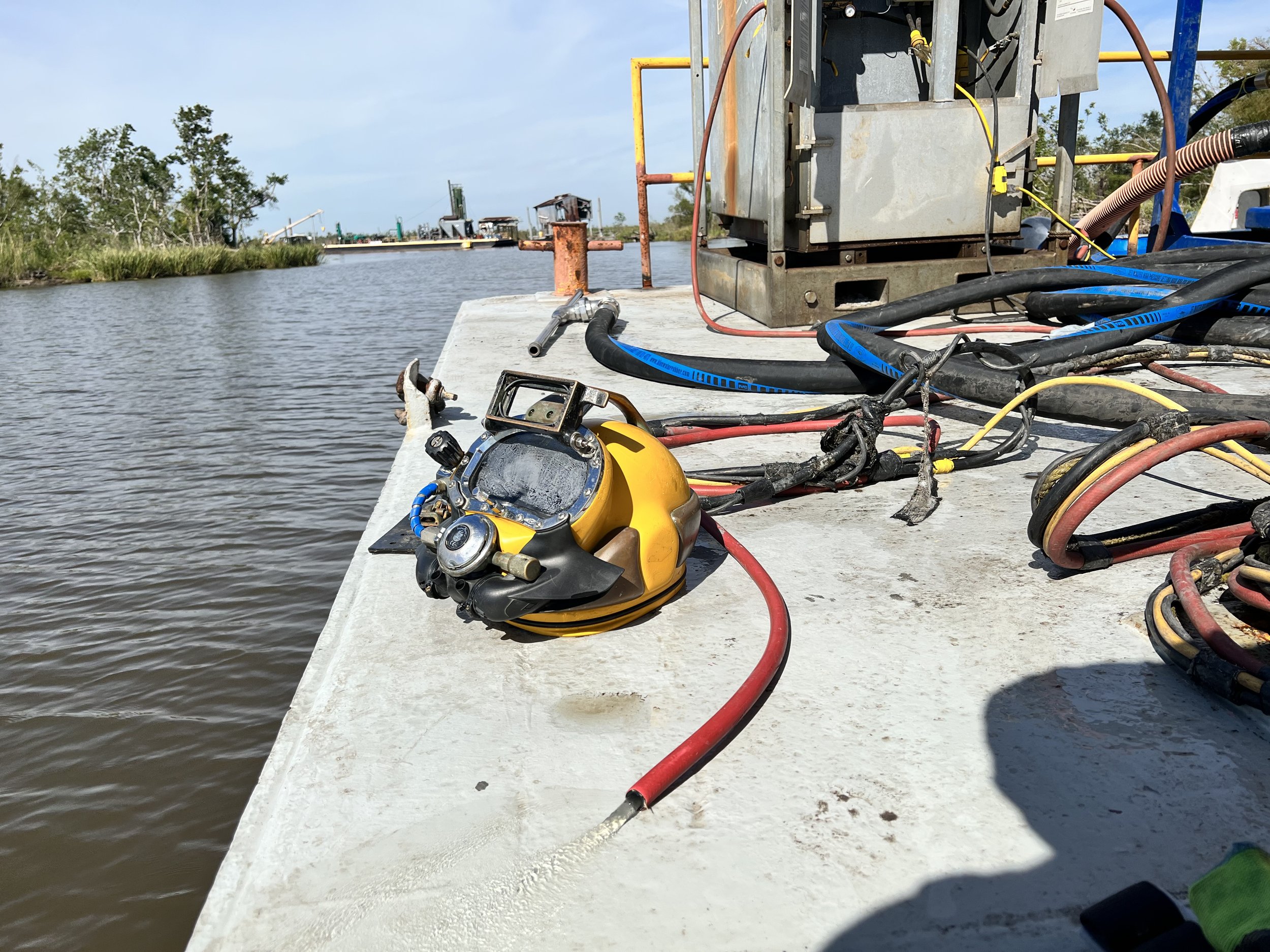 A yellow diving helmet with a transparent face shield and attached hoses is resting on a concrete dock beside several electrical cables and equipment near a body of water.