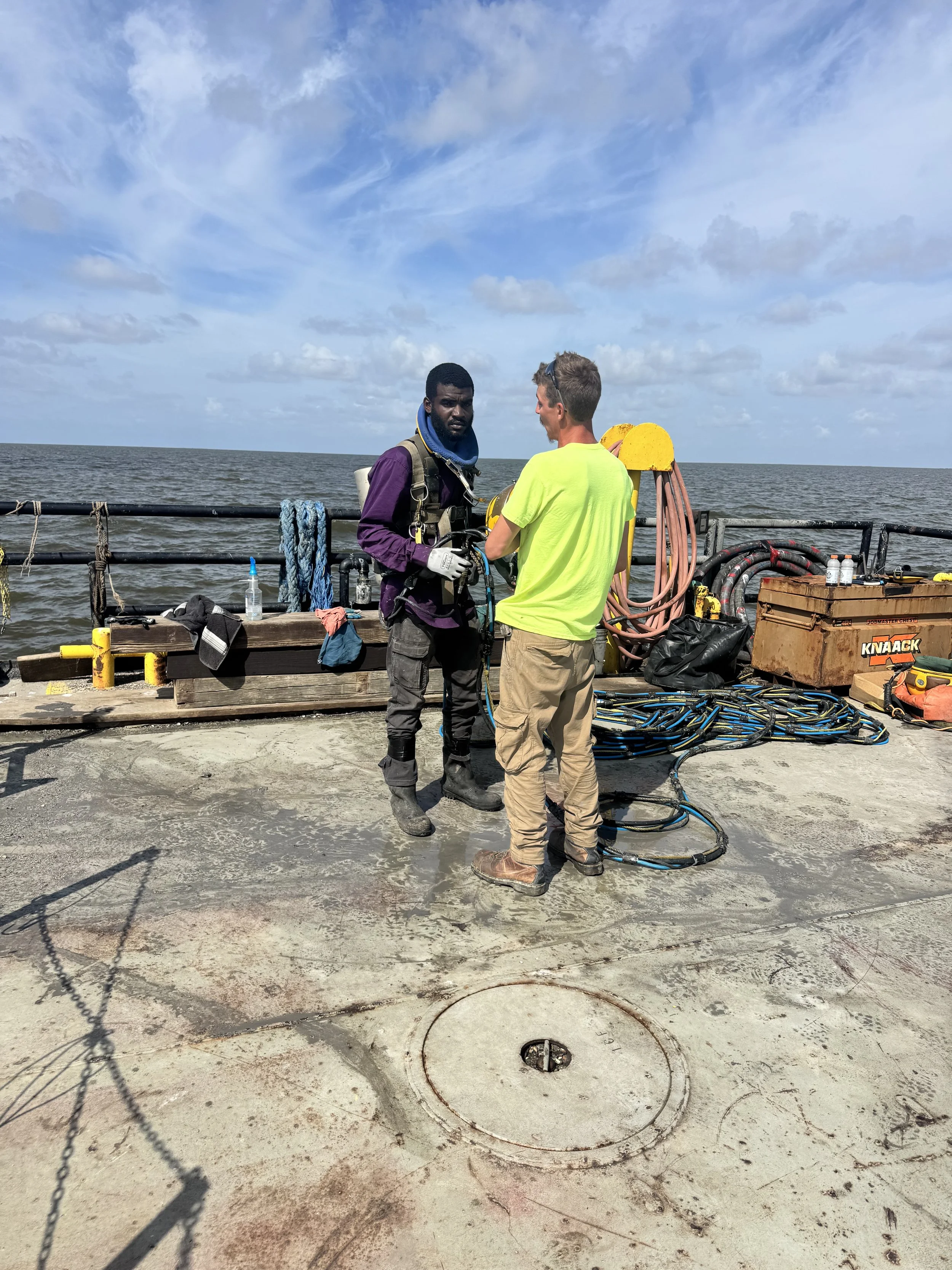 Two men on a boat deck, one in a purple and black suit with safety gear, the other in a neon yellow shirt and beige pants, preparing for a task near the ocean with ropes, tools, and equipment surrounding them.