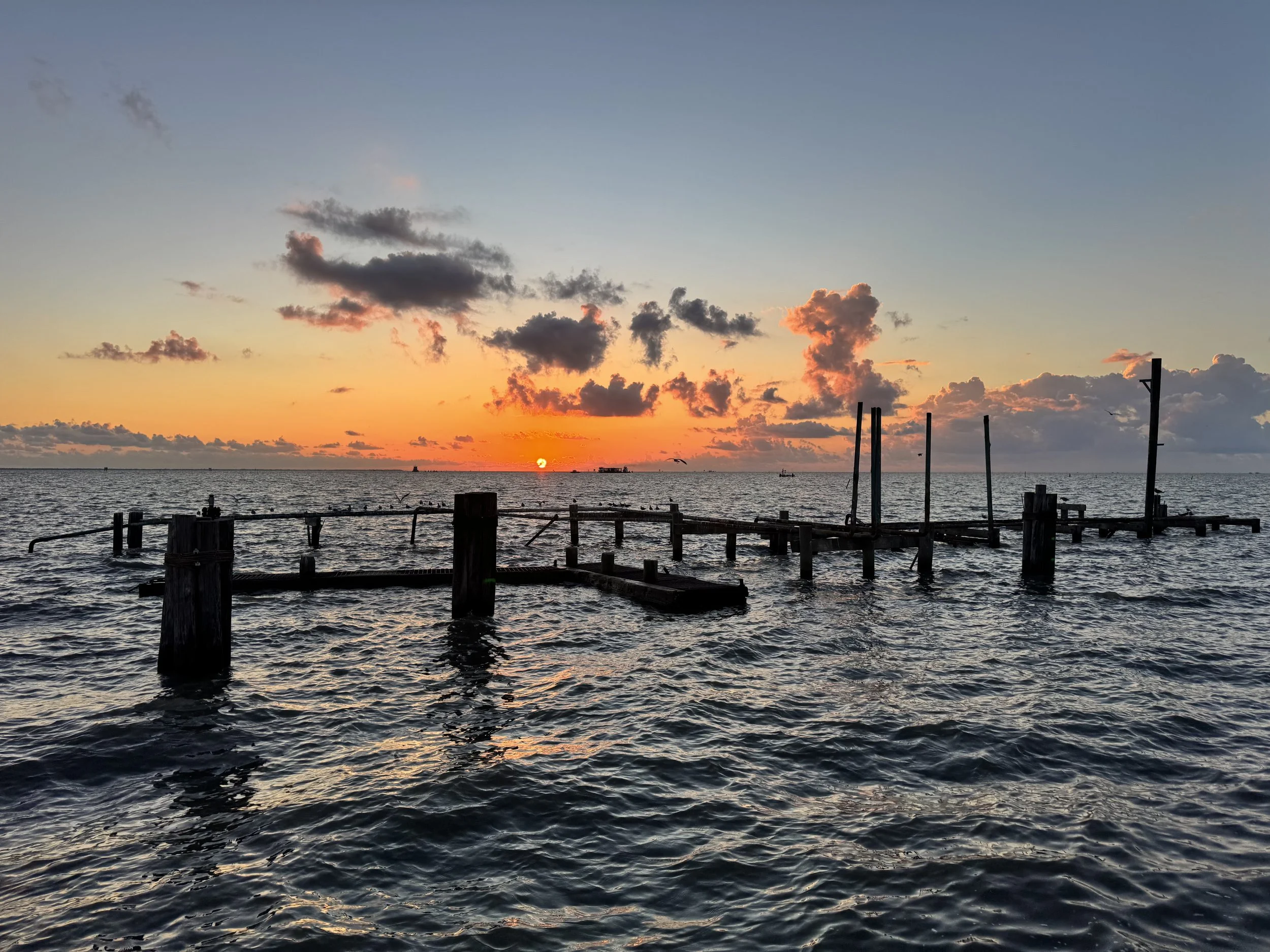 Sunset over the ocean with a partially submerged wooden pier and boats in the distance.