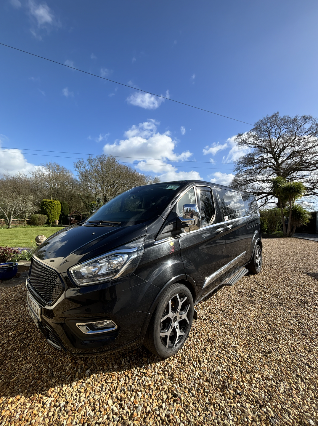 Black van parked on a gravel driveway under blue sky with scattered clouds, surrounded by trees and lawn.