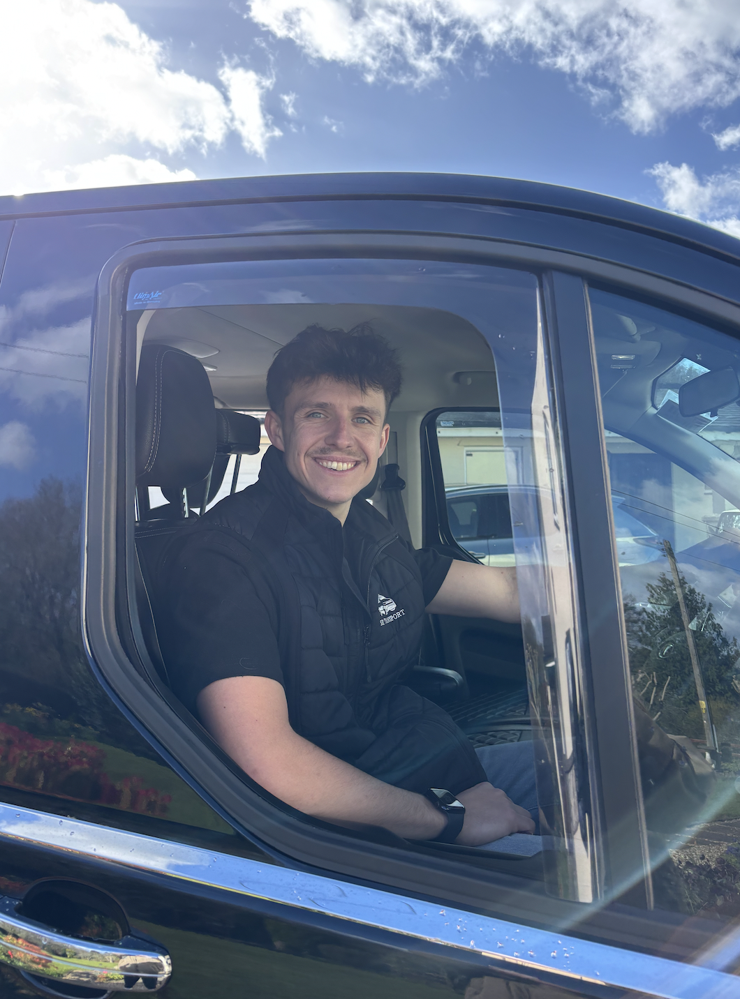 A smiling young man sitting in the driver's seat of a black vehicle, looking out the open window, with a partly cloudy sky overhead.