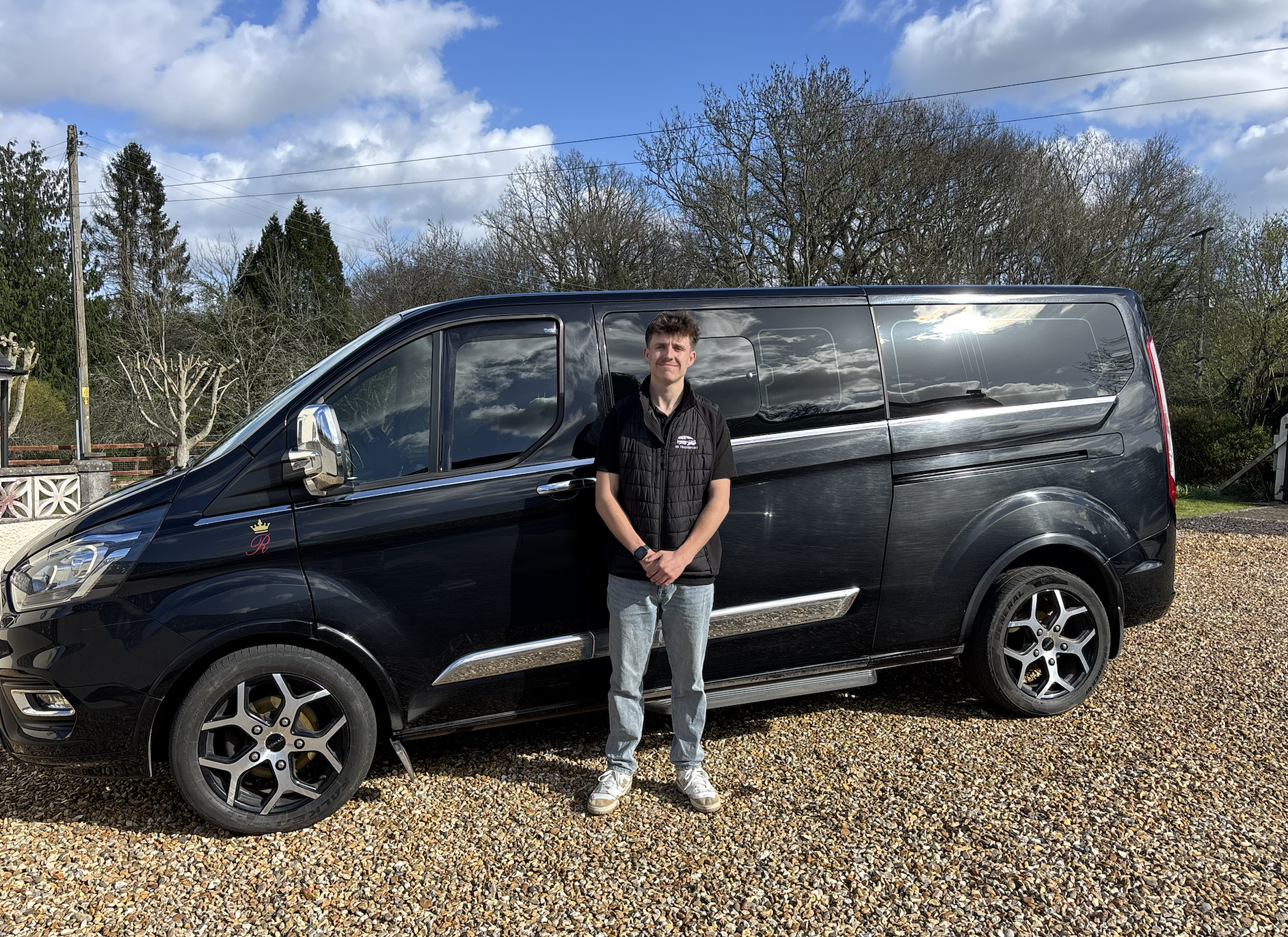 A young man stands in front of a black van on gravel with trees and a partly cloudy sky in the background.