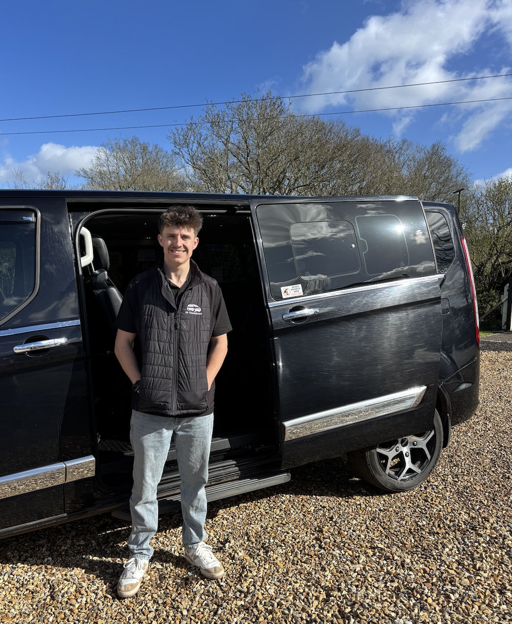 A young man standing next to a black van with the sliding door open, on a gravel surface with trees and a blue sky with clouds in the background.