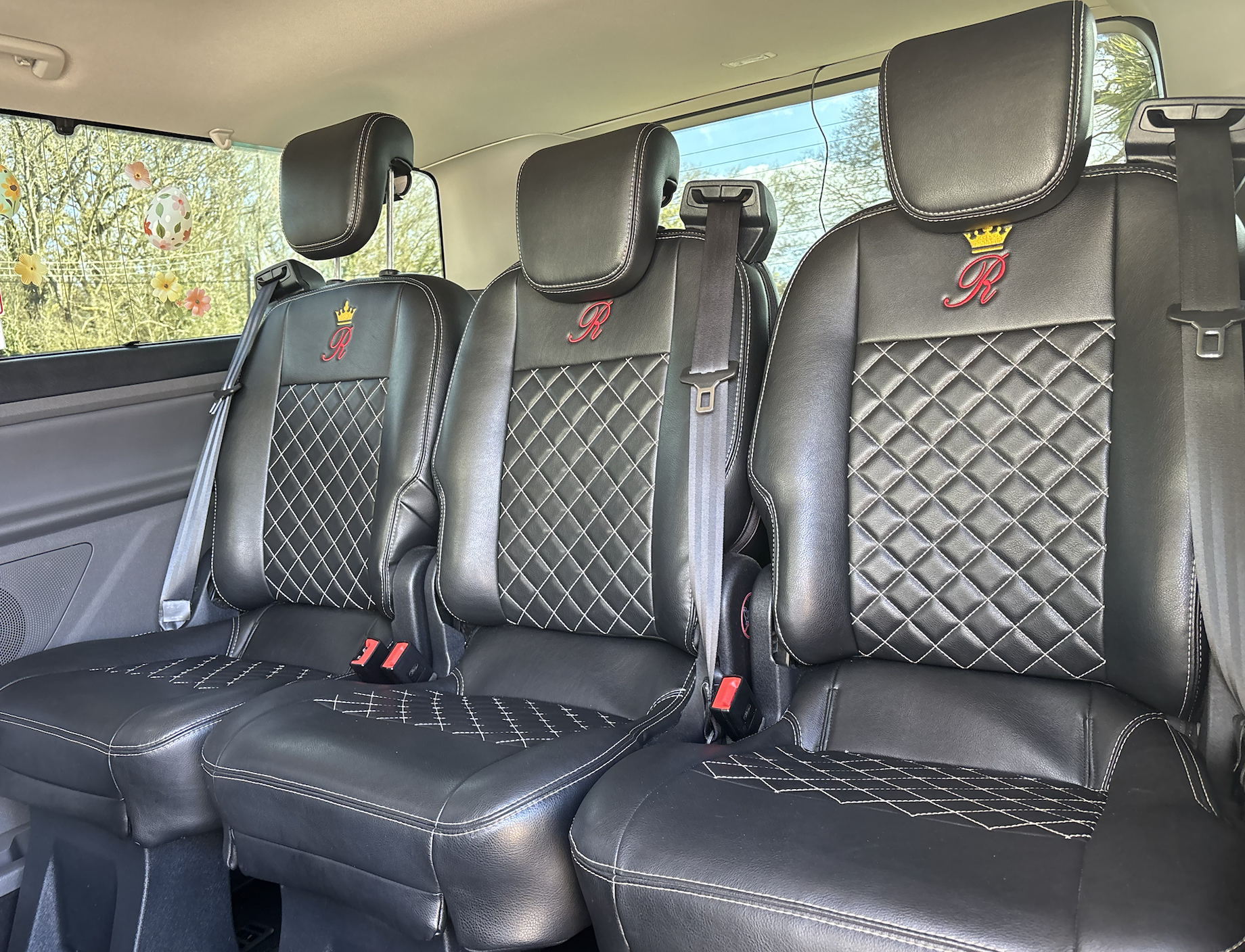 Interior of a vehicle showing three black leather seats with embroidered red letter 'R' and a yellow crown above it, featuring diamond-stitched patterns and headrests, with a window decorated with Easter egg and flower stickers visible in the background.