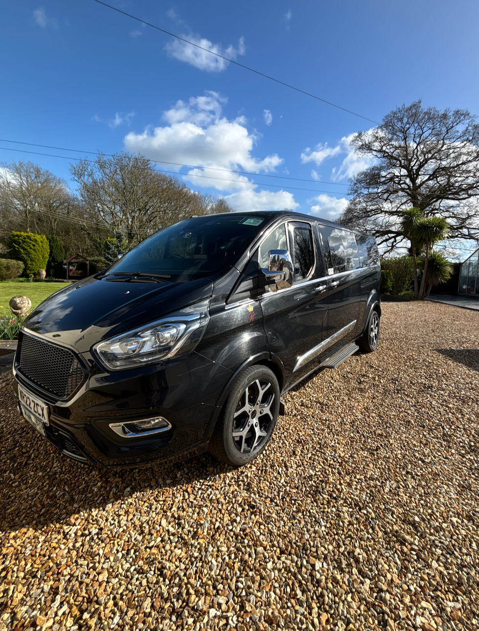 Black van parked on a gravel driveway with a garden and trees in the background under a blue sky with clouds.