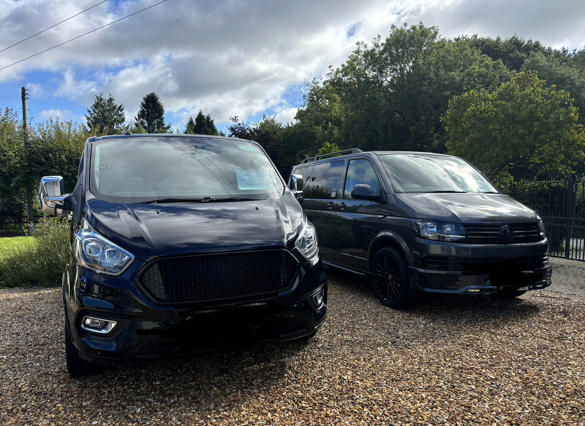 Two black vehicles, a van on the left and a minivan on the right, parked on a gravel lot with trees and a cloudy sky in the background.