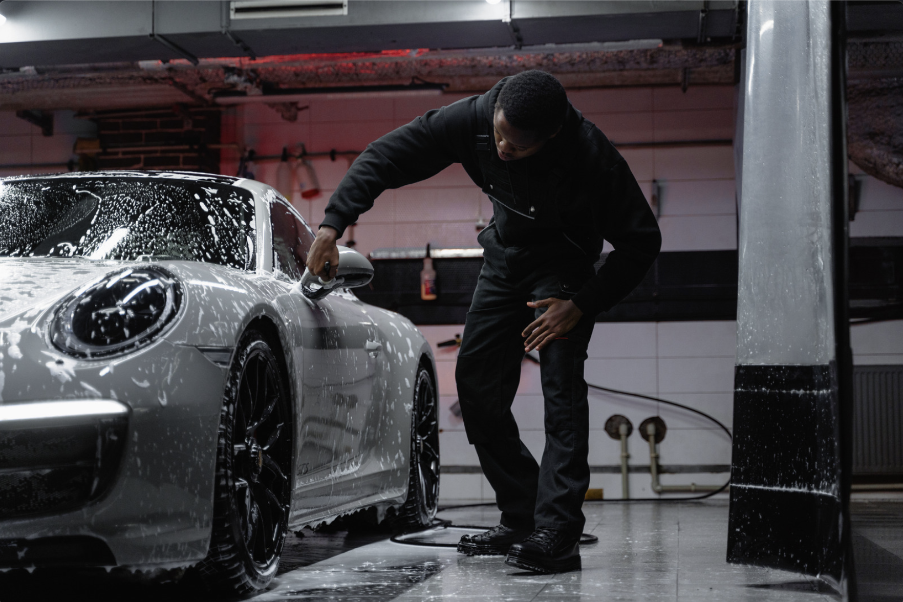 Man washing a silver sports car with soap suds in a garage.