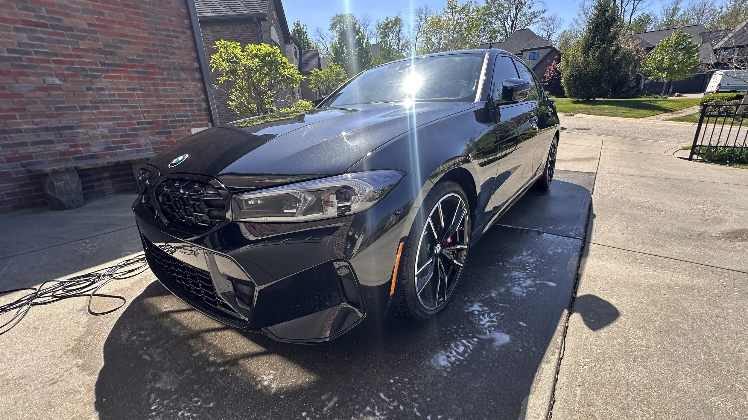 Black BMW car parked on a driveway on a sunny day with clear sky. The driveway has some soap suds and a hose nearby. Houses with brick walls and trees can be seen in the background.