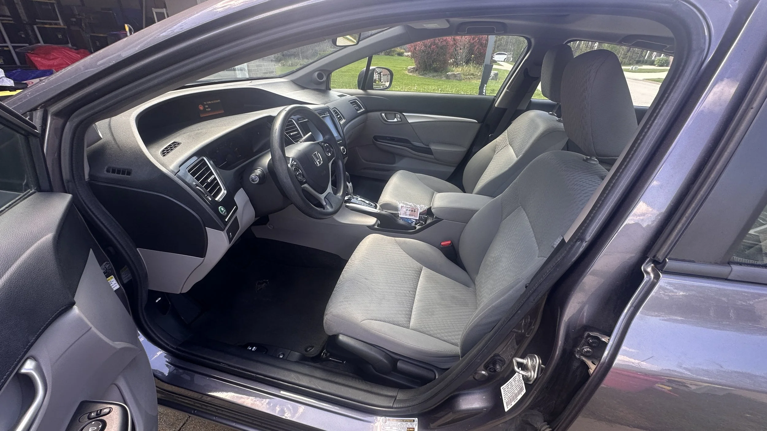Interior of a gray Honda car showing front seats, dashboard, steering wheel, and gear shift, with a glimpse of the outside green lawn and trees through the windows.