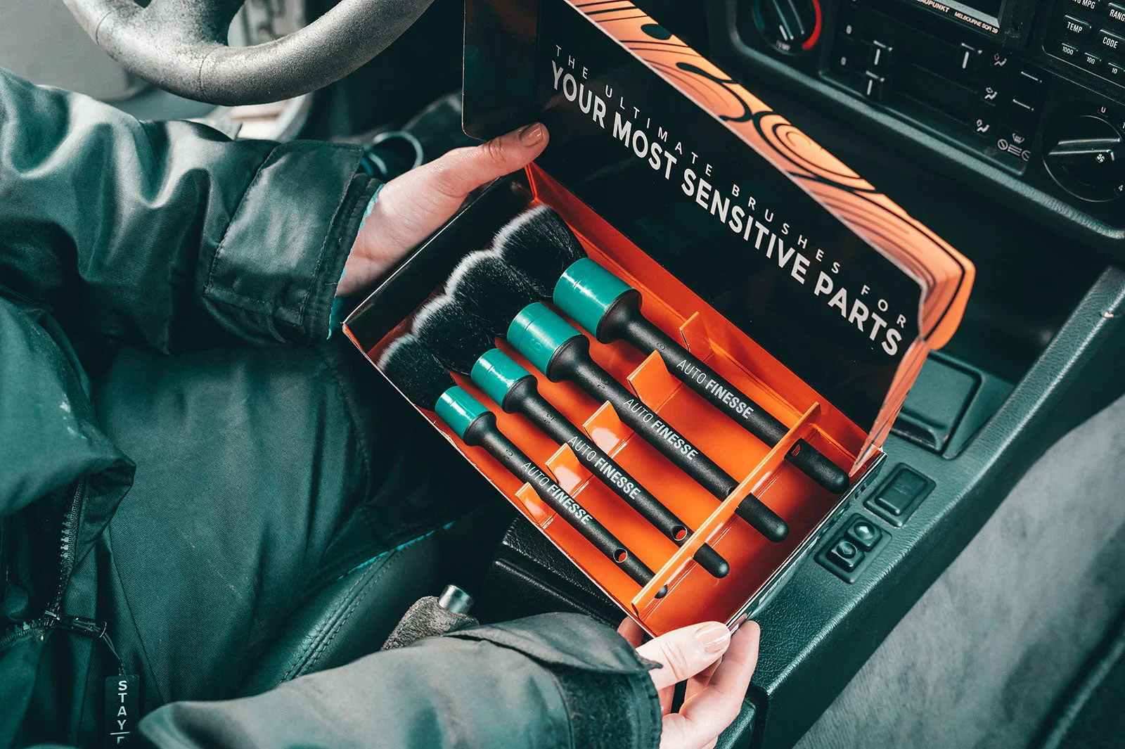 Person holding a box of auto brushes labeled 'auto finesse' inside a vehicle, with the vehicle's dashboard and steering wheel visible.