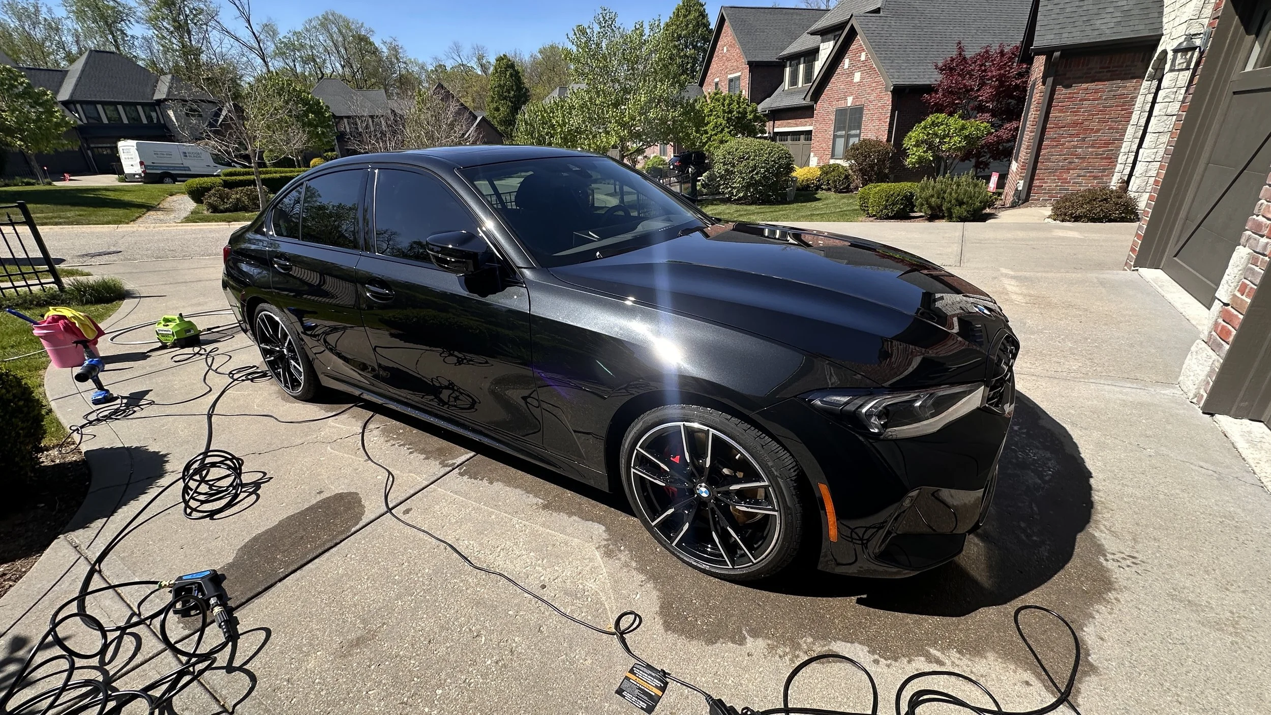 Black sports car in a driveway with car washing equipment and extension cords scattered around.