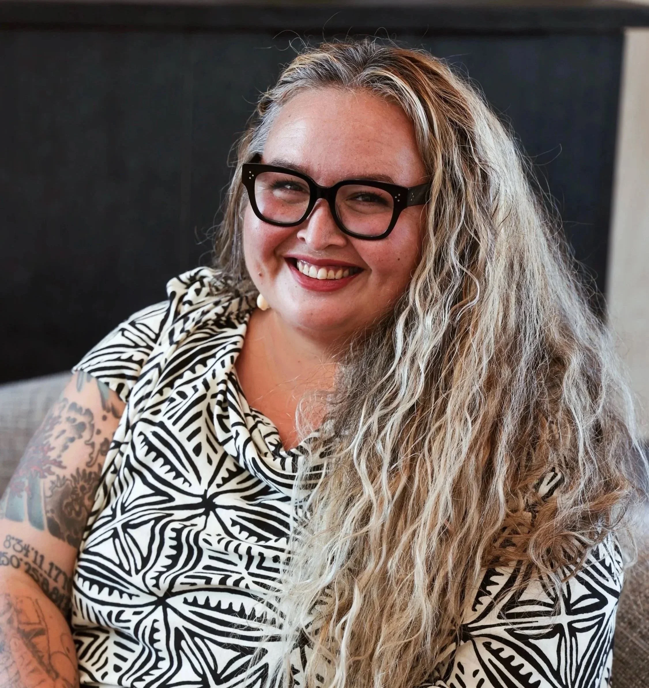 A smiling woman with curly blonde hair, wearing black glasses and a black and white patterned top, sitting indoors.