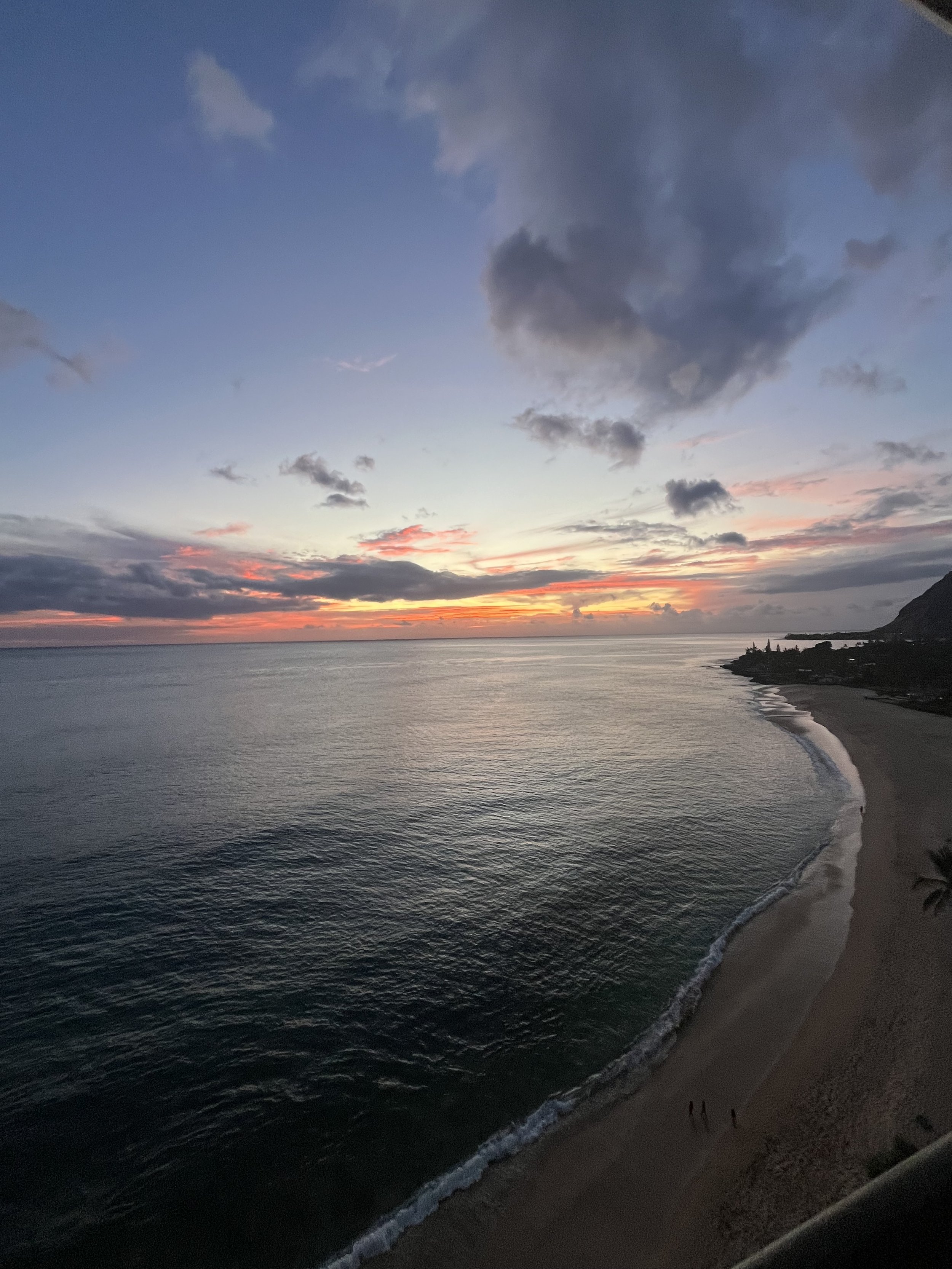 Sunset over the ocean with a partly cloudy sky, a sandy beach, and some small figures walking along the shoreline.