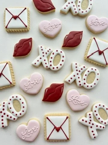 Decorative cookies shaped like lips, hearts with 'Love' written on them, envelopes with red ribbon, and 'XOXO' messages with red polka dots.