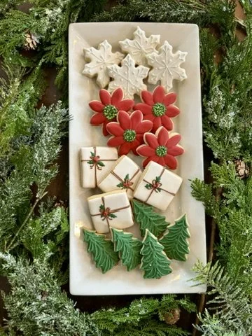 Assorted Christmas cookies on a rectangular white plate, decorated as poinsettias, holly leaves, and small gift boxes, surrounded by greenery.