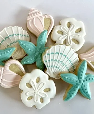 Assorted decorated sugar cookies shaped like shells, starfish, and flowers, decorated with pastel icing in pink, white, blue, and beige.