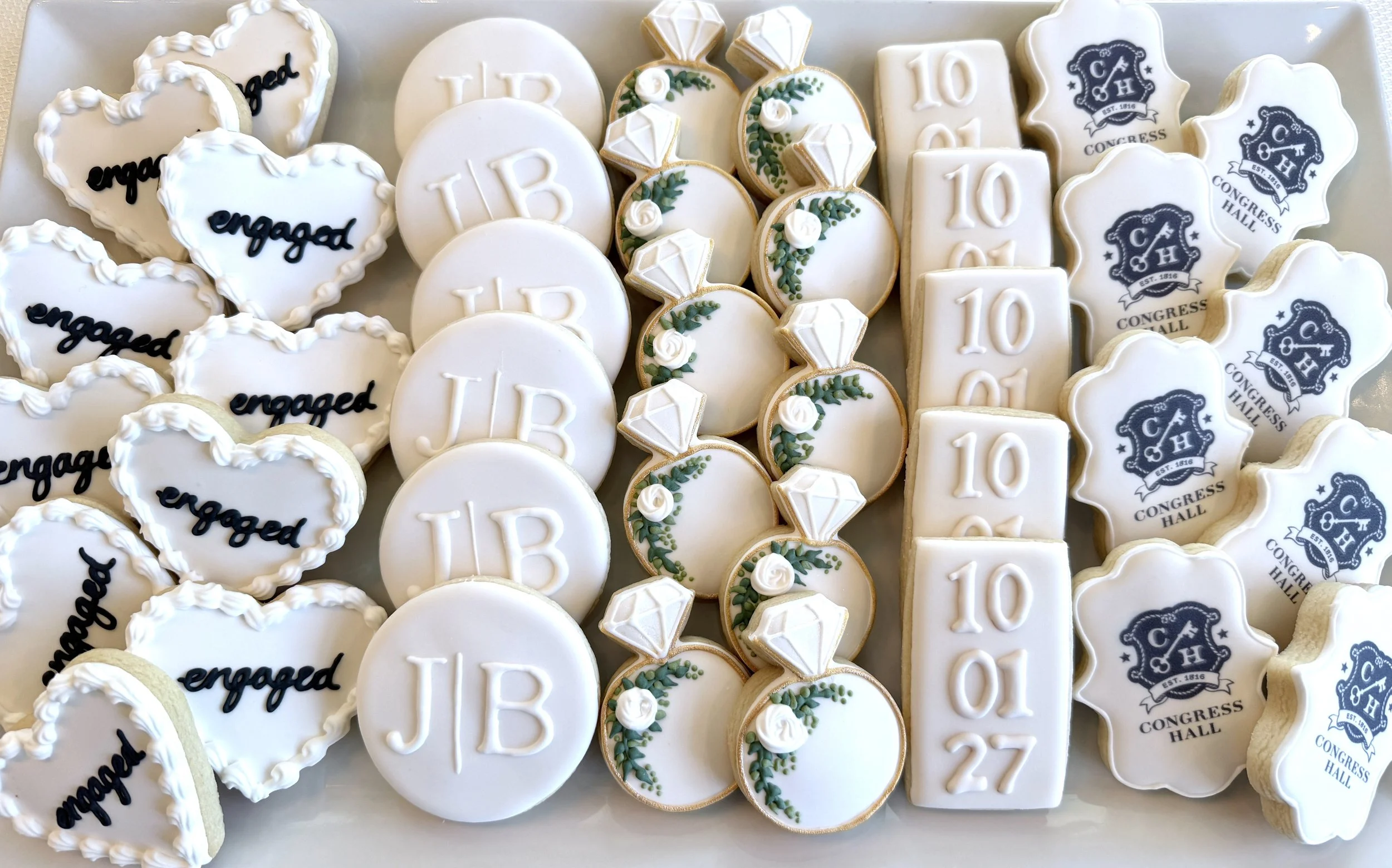 Assorted decorated cookies, including heart-shaped with 'engaged' written, round with 'I B', rectangular with '10 06', oval with green leaves and white flowers, and logo cookies for Congress Hall with navy blue emblem.