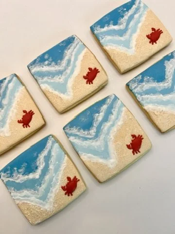 Five beach-themed decorated sugar cookies with blue ocean waves, sandy shores, and a red crab on each.