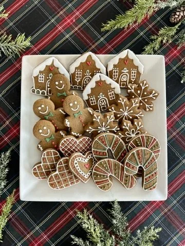 Assorted decorated Christmas cookies on a white square plate, including gingerbread men, house-shaped cookies, snowflakes, mittens, and peppermint candies, with pine branches around the plate on a plaid tablecloth.