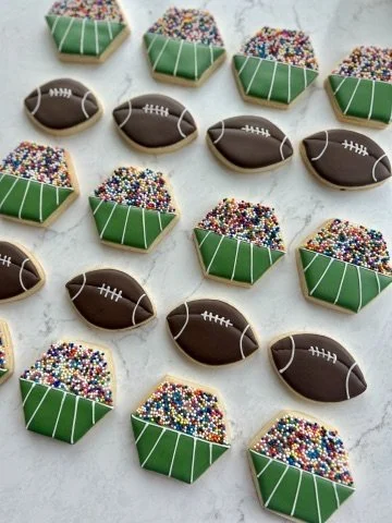 Decorated cookies shaped like footballs, stacks of colorful sprinkles, and football stadiums with green grass and white lines, arranged on a light-colored surface.