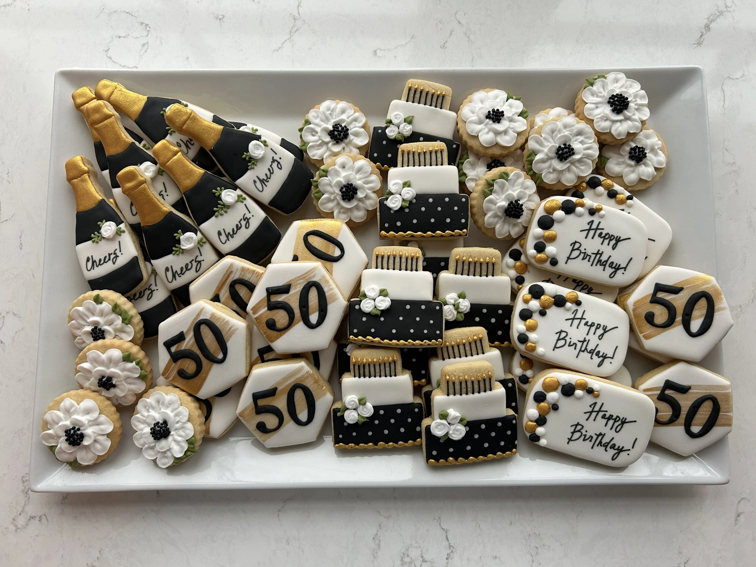 A white rectangular tray filled with decorated cookies for a birthday celebration. The cookies include champagne bottles with black, white, and gold details, birthday cakes with black, white, and gold decorations, and flowers with white petals and bl