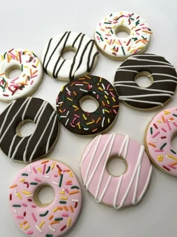 An assortment of colorful donuts with pink, white, and chocolate frosting, topped with rainbow sprinkles and decorative stripes, arranged on a white surface.