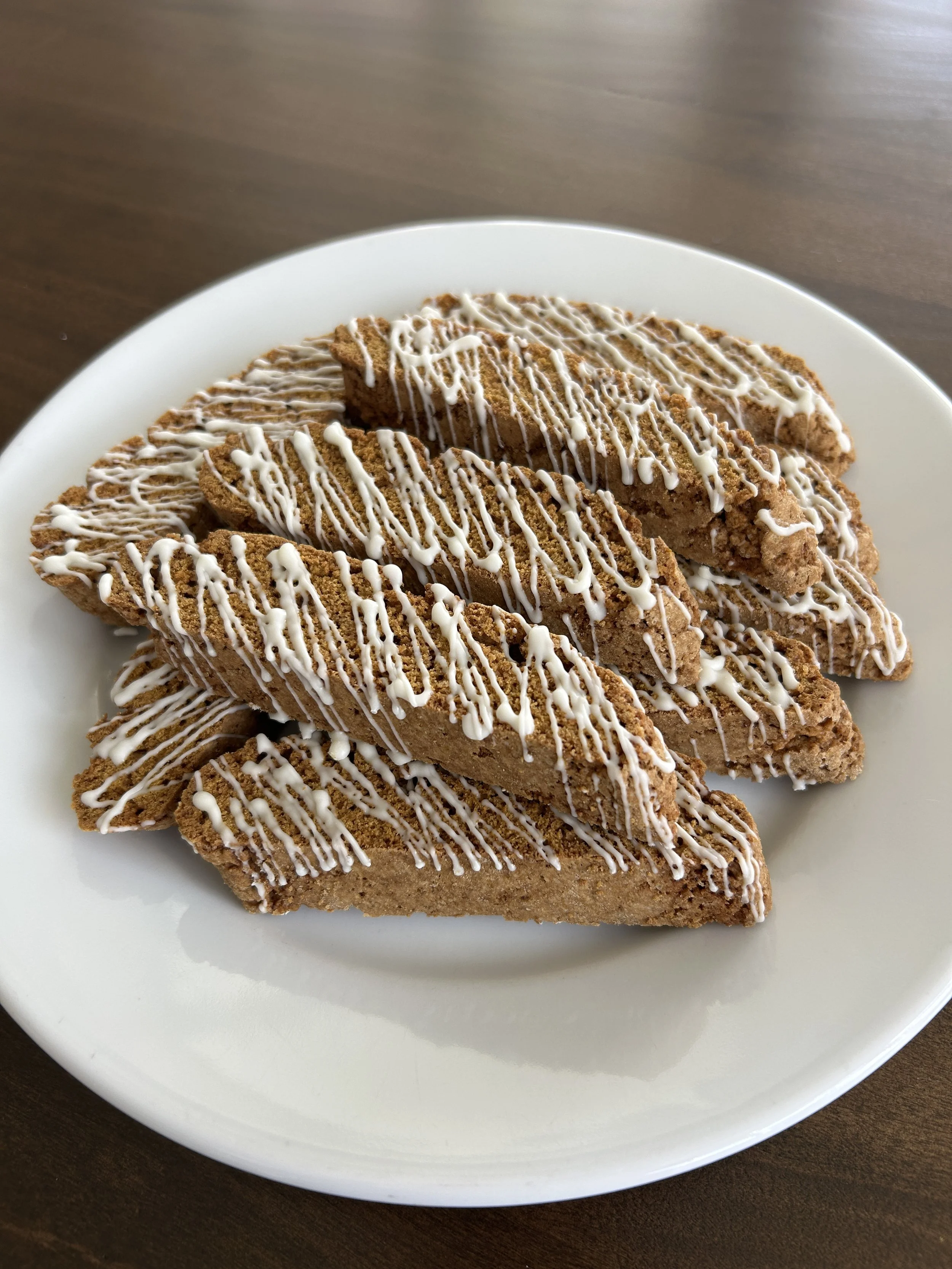 Three pieces of biscotti with white chocolate drizzle on a white plate, placed on a dark wooden surface.