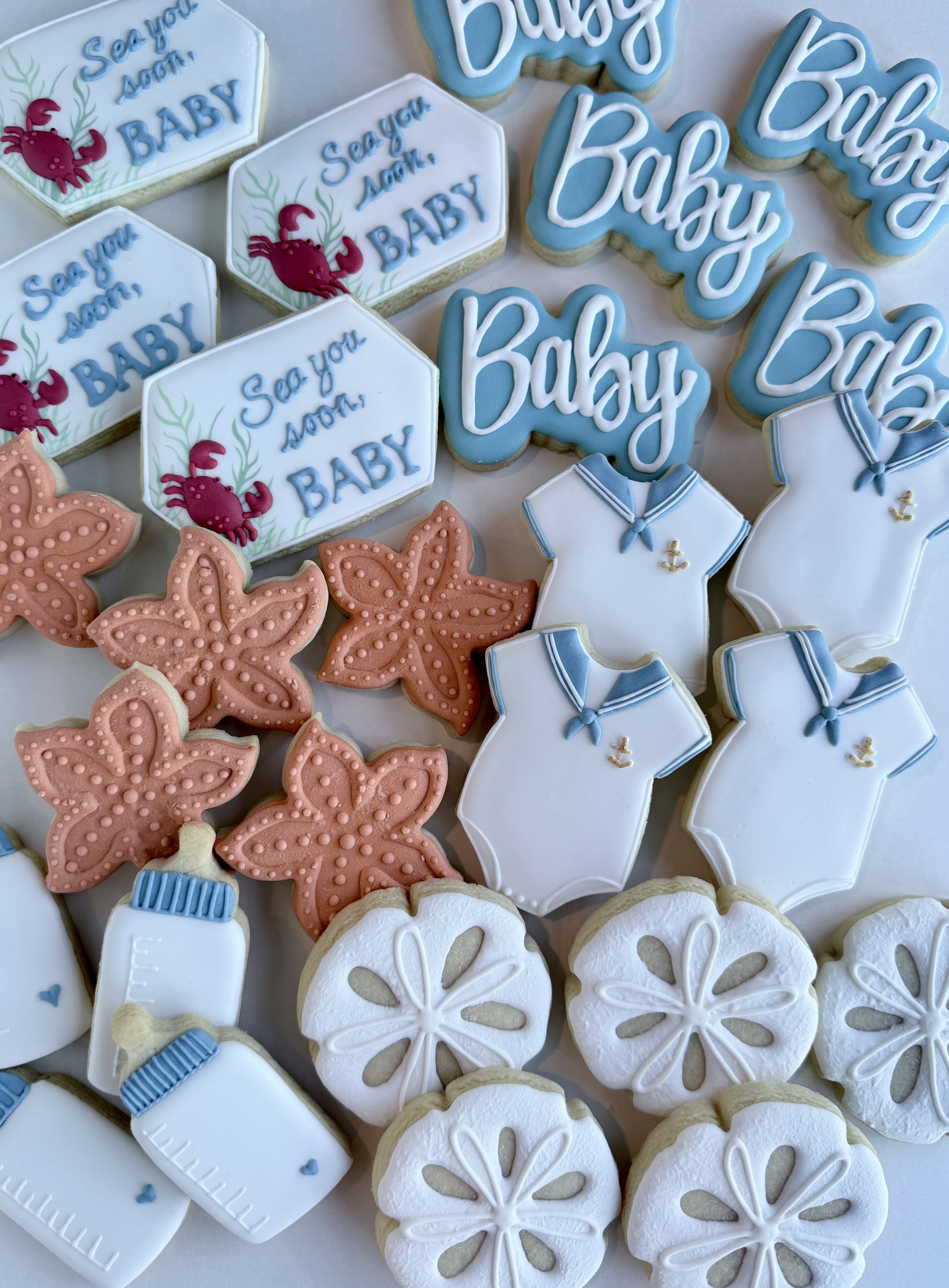 Cookies decorated for a baby shower, including ones with the phrase "Say You Soon Baby" with a red crab illustration, ones resembling baby onesies with blue details, and round cookies with a white flower pattern, some with peach-colored leaves.
