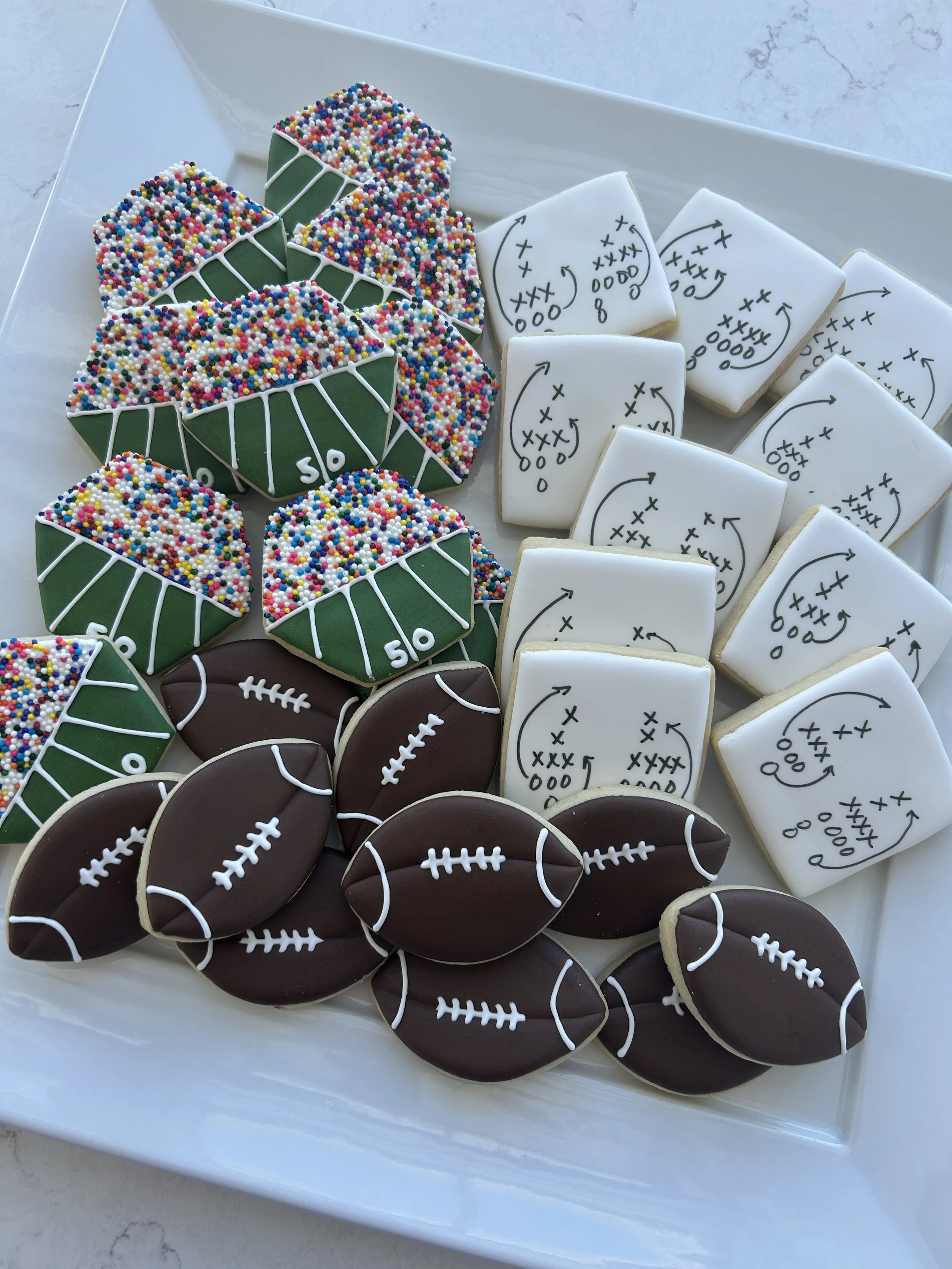 Assorted football-themed cookies, including decorated sugar cookies with white icing and football outlines, and cookies decorated with football field diagrams and colorful sprinkles, arranged on a white tray.