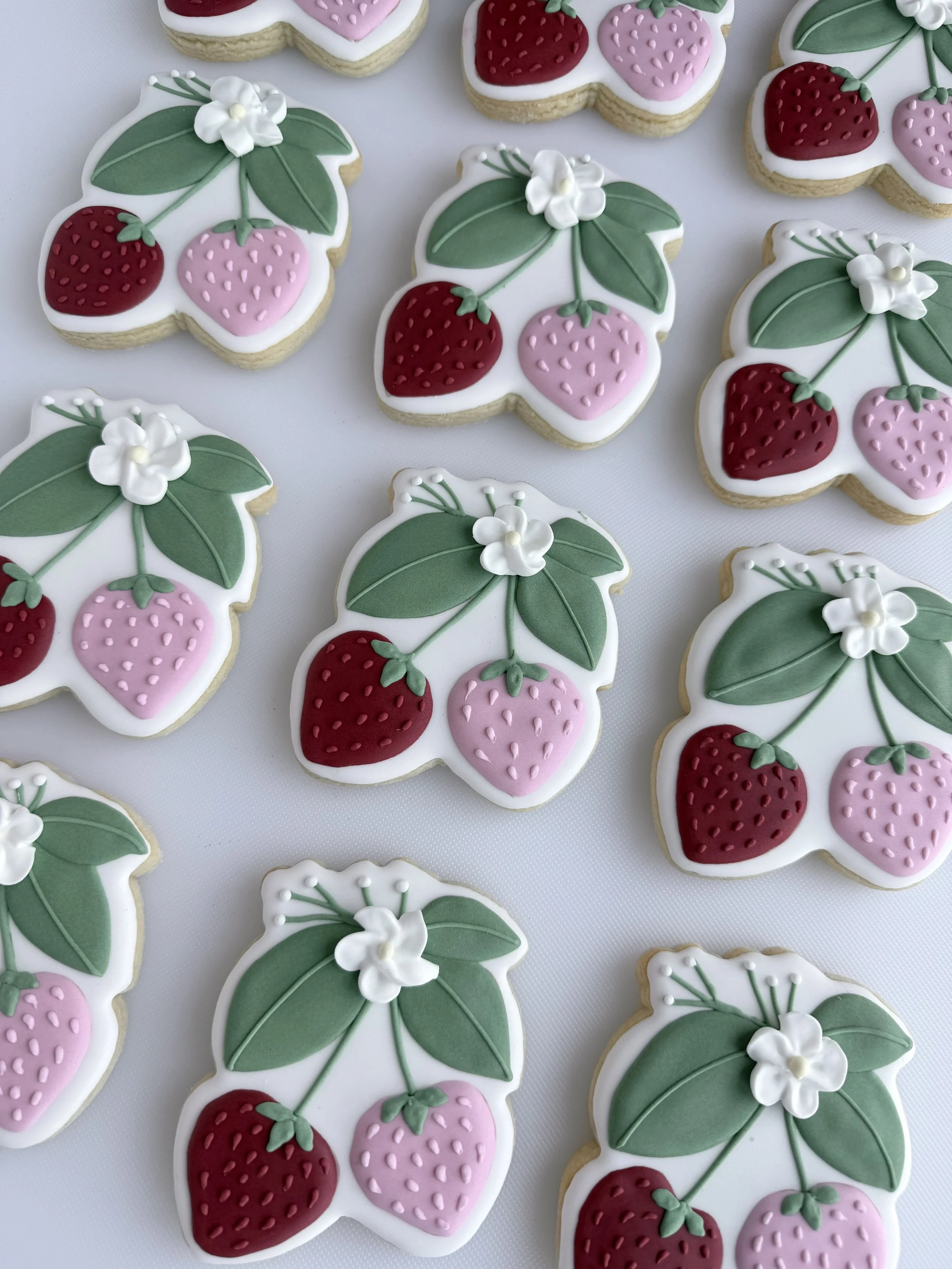 Decorated cookies shaped and decorated like strawberries and leaves, with white icing and small white flowers.