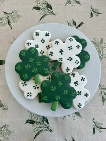 Plate of decorated cookies shaped like shamrocks, with green and white icing, on a floral tablecloth.