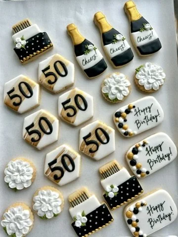 Birthday cake cookies shaped like champagne bottles, birthday hats, and flowers, with some cookies decorated with the number 50 and others with 'Happy Birthday!', arranged for a celebration.