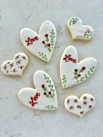 Six decorated sugar cookies in heart shapes with red and green floral designs and the word "Love" on some.