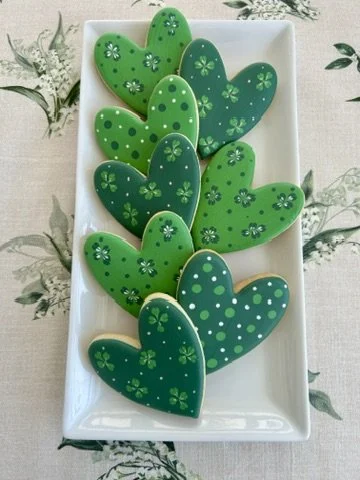 A rectangular white plate holding eight green heart-shaped cookies decorated with various floral and polka dot patterns.
