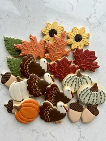 Assorted fall-themed cookies shaped like sunflowers, leaves, pumpkins, turkeys, and gourds on a white surface.