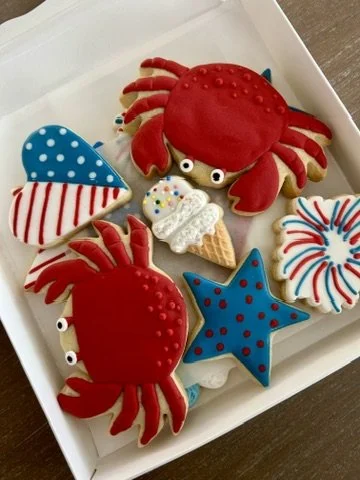 Decorated cookies in a box including red crabs, a crab with brown eyes, a blue star, a blue semi-circle with white dots, a white circular cookie with red, blue, and yellow sprinkles, and a white circular swirl cookie with red and blue lines.