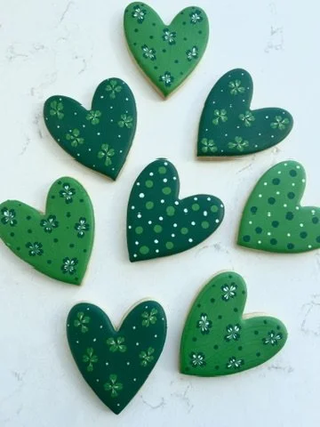 Nine green heart-shaped cookies decorated with various small flower and dot patterns on a light background.