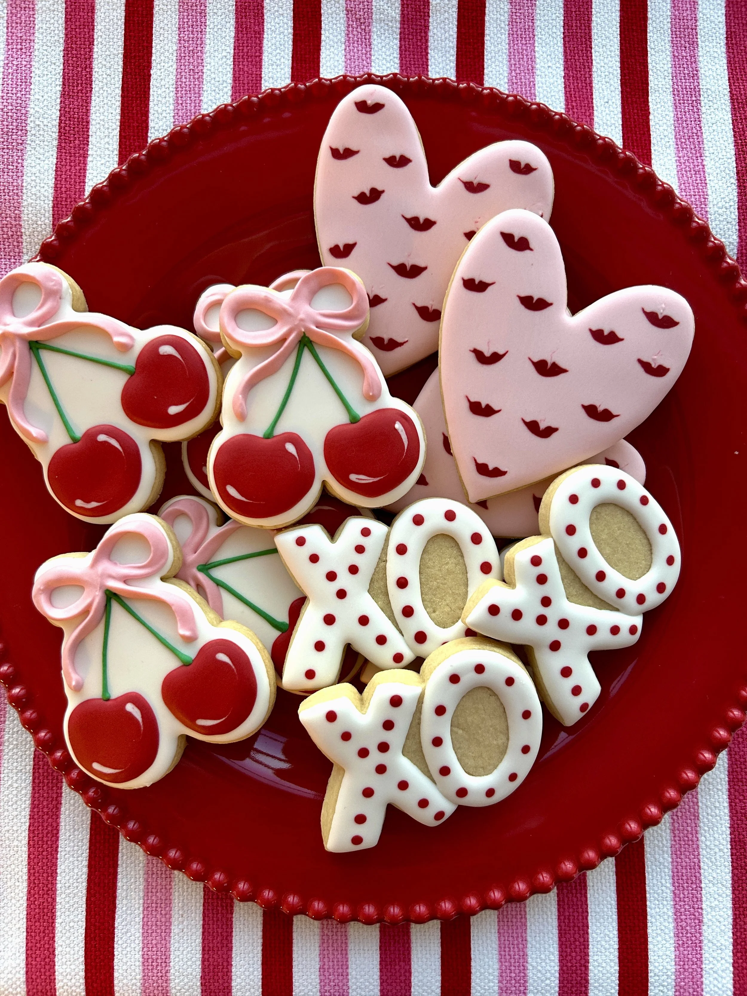 Cookies decorated with cherries, lips, and hearts, arranged on a red plate with a striped tablecloth background.