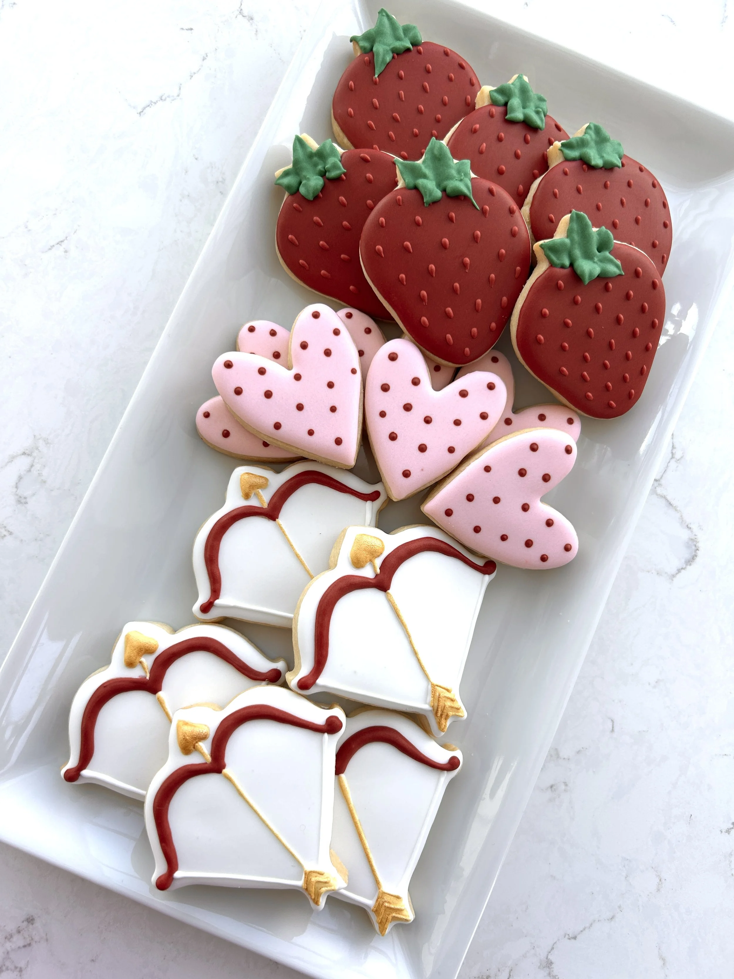 Decorated cookies on a white platter, shaped like strawberries, hearts, and flamingos, with red, pink, green, and white icing.