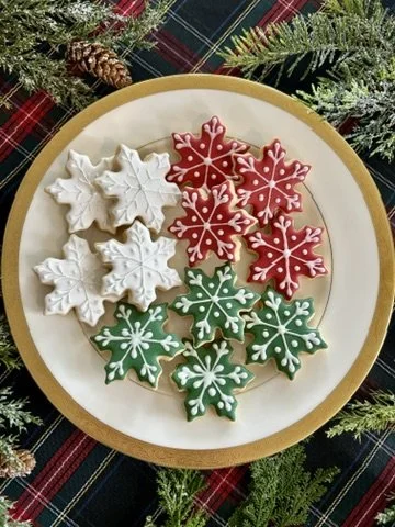 Plate with Christmas-themed decorated cookies shaped like snowflakes, in red, white, and green, on a festive table with pine branches and pinecones.