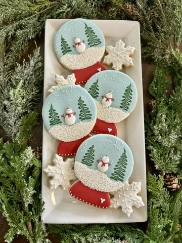 Holiday cookies on a white tray surrounded by pine branches. The cookies include three decorated with winter trees and snowmen, and two shaped like snowflakes.