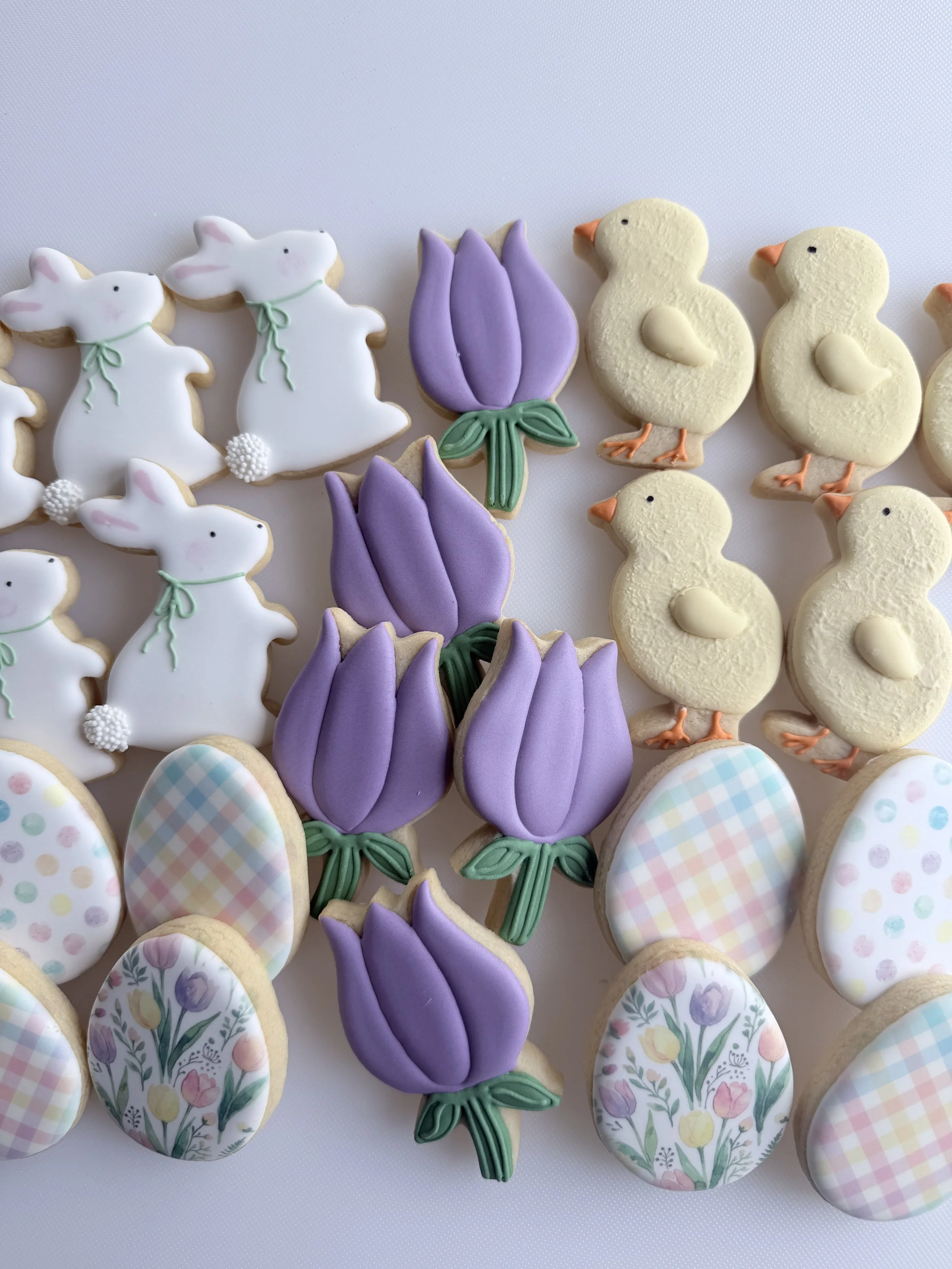 Decorated cookies shaped like bunnies, purple tulips, ducklings, and decorated eggs for Easter, arranged on a white surface.